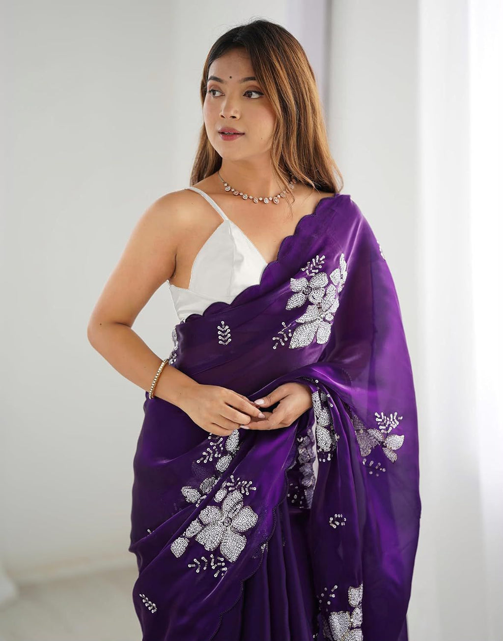 A woman modeling a purple saree with silver floral embroidery, paired with a white sleeveless blouse, set against a bright, neutral indoor background.