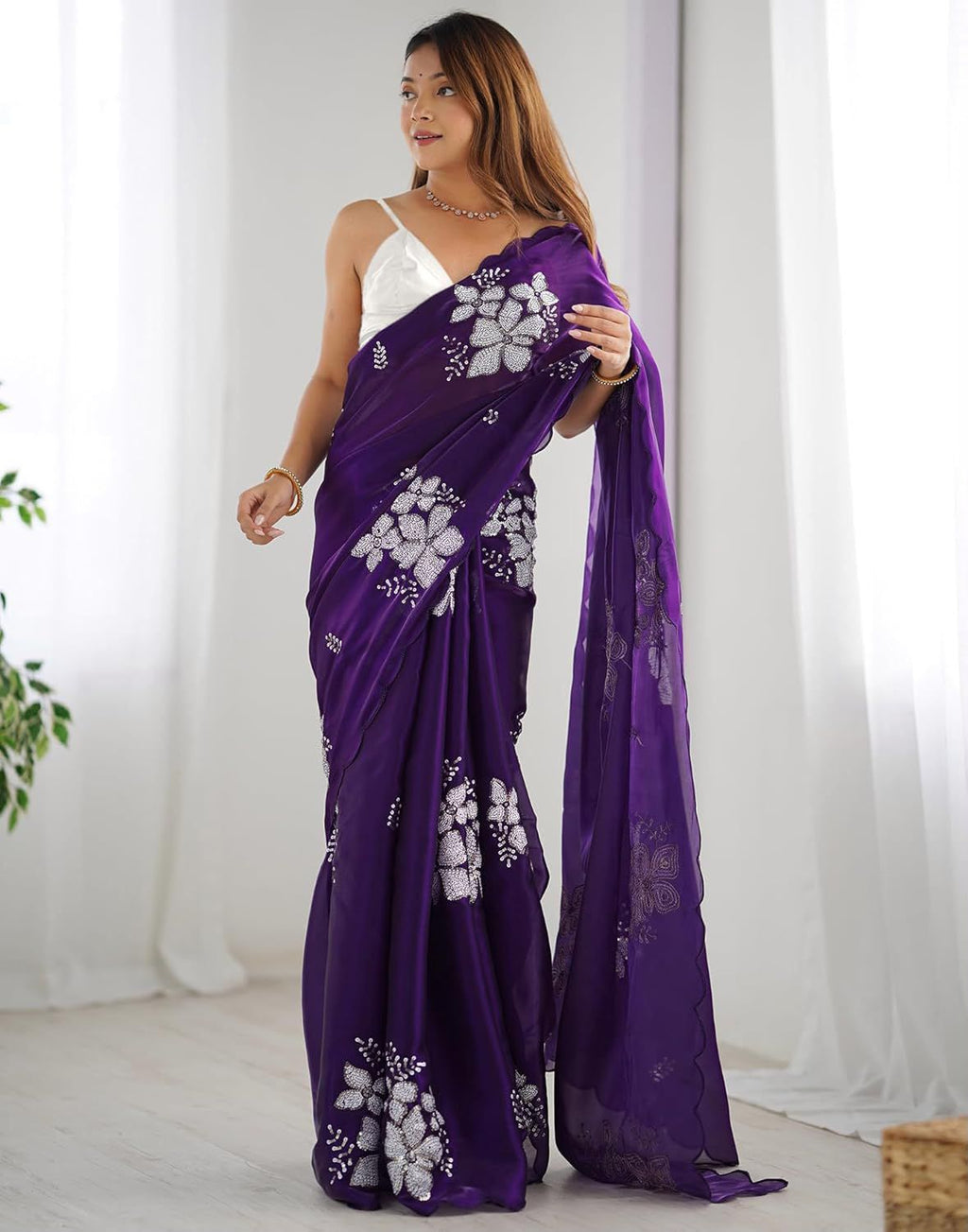 A woman models a purple saree with white floral prints and a white sleeveless blouse in a bright, minimalist room with white curtains.