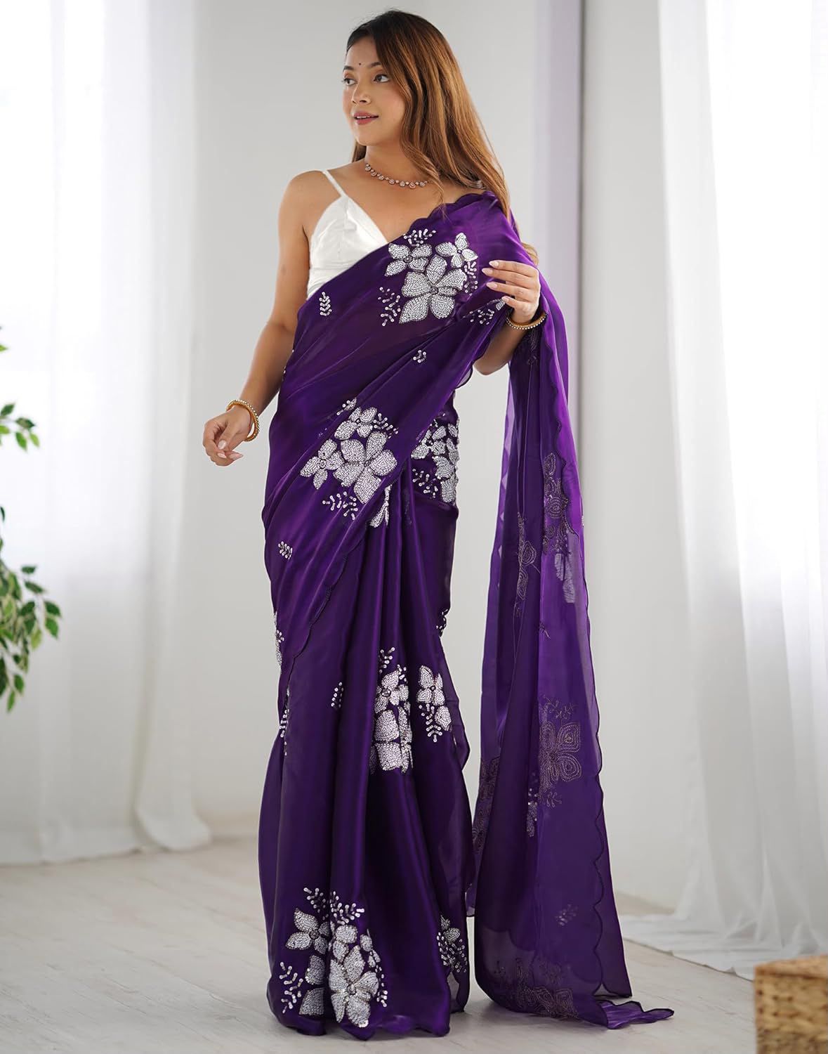 A woman models a purple saree with white floral prints and a white sleeveless blouse in a bright, minimalist room with white curtains.